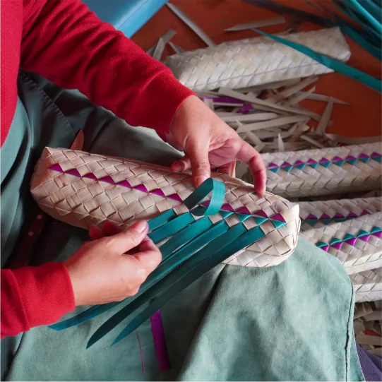 A woman is skilfully peeling a banana stem to prepare fibres for craft weaving. She is seated at a workstation, using a knife or similar tool to carefully remove the outer layers of the stem. The peeled fibres are laid out neatly beside her, ready to be dried and woven into intricate crafts. The setting is a traditional or artisanal workshop, emphasising the meticulous and hands-on nature of the craft.