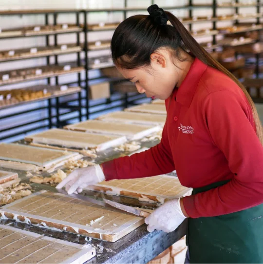 A woman is carefully handling a tray of soap in the workshop. She is wearing gloves and working at a table with multiple trays in the background, indicating a handmade, artisanal production process for natural cosmetics.