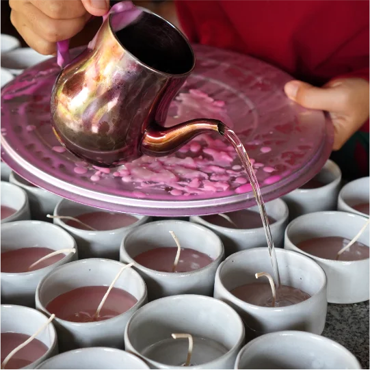 A woman is carefully trimming wicks on handmade candles in a workshop. The candles, arranged in neat rows on a table, appear to be freshly made, with trimmings scattered on the surface, indicating an artisanal production process focused on natural scents and quality craftsmanship.